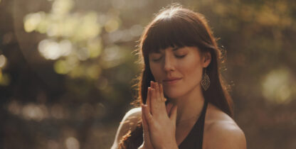 Brown haired woman with eyes closed and hands in a prayer position outside in dabbled sunlight